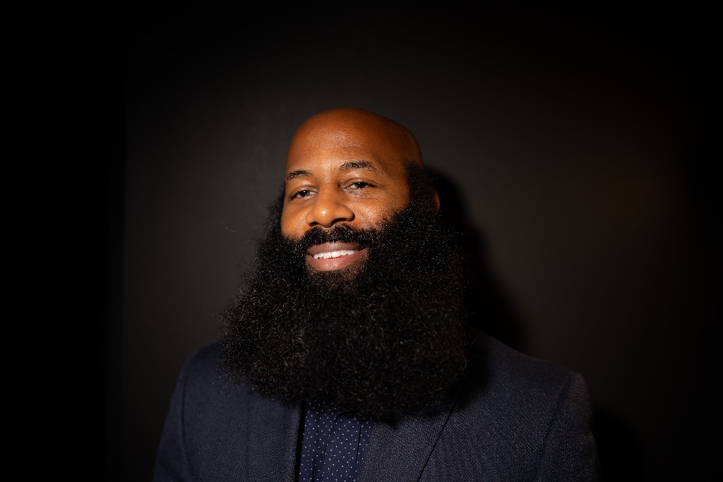 Studio portrait of Anson smiling warmly in a tailored dark navy jacket and patterned shirt, with his full beard and natural studio lighting against a dark charcoal backdrop.